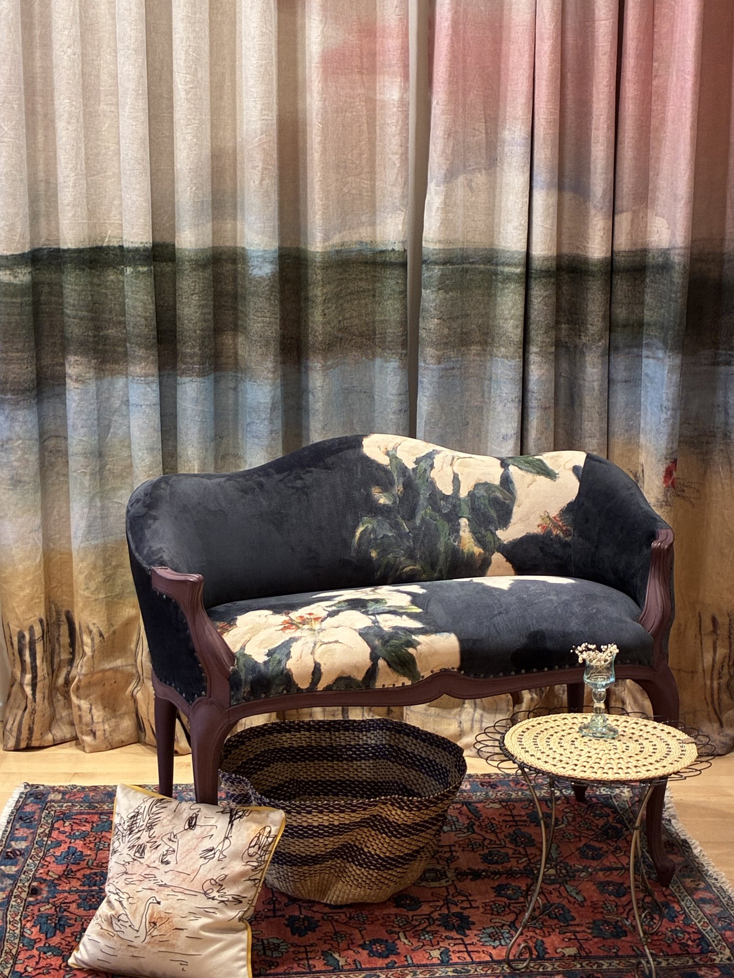 Painterly upholstered settee, side table and pillows in front of landscape-print curtains